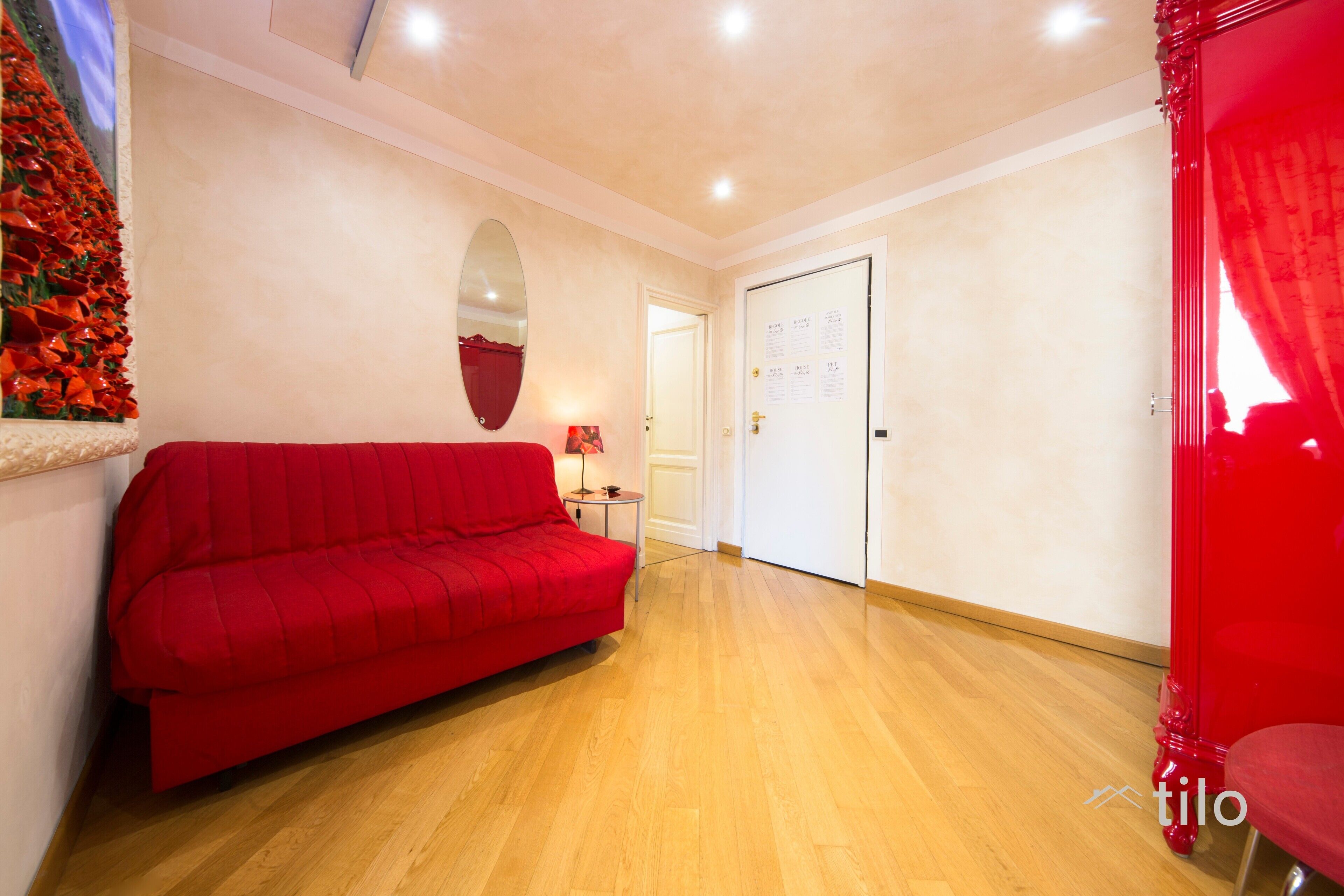 Une pièce avec un canapé rouge, une petite table avec une lampe, une porte sur laquelle est fixé un tableau d’affichage, et un miroir au mur.