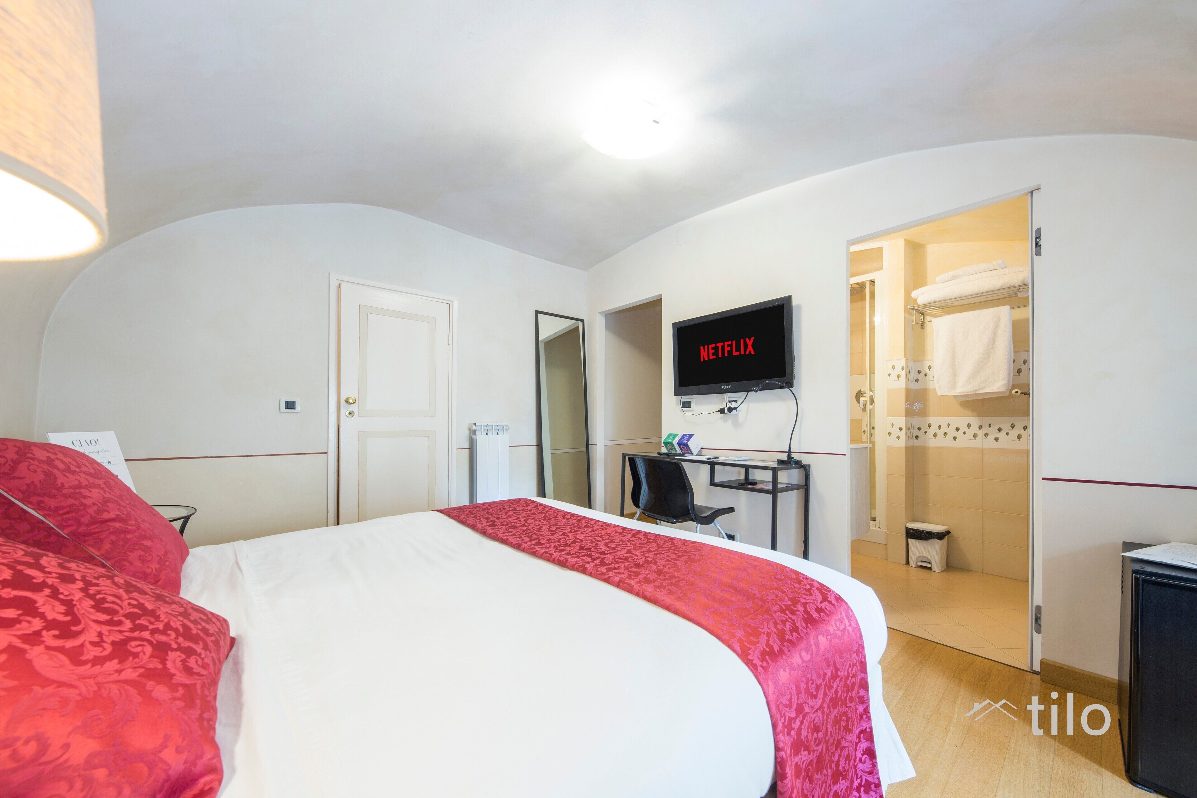 Une chambre d’hôtel avec un lit, un bureau équipé d’un ordinateur, un téléviseur affichant le logo « NETFLIX », et une salle de bain visible à travers une porte ouverte.