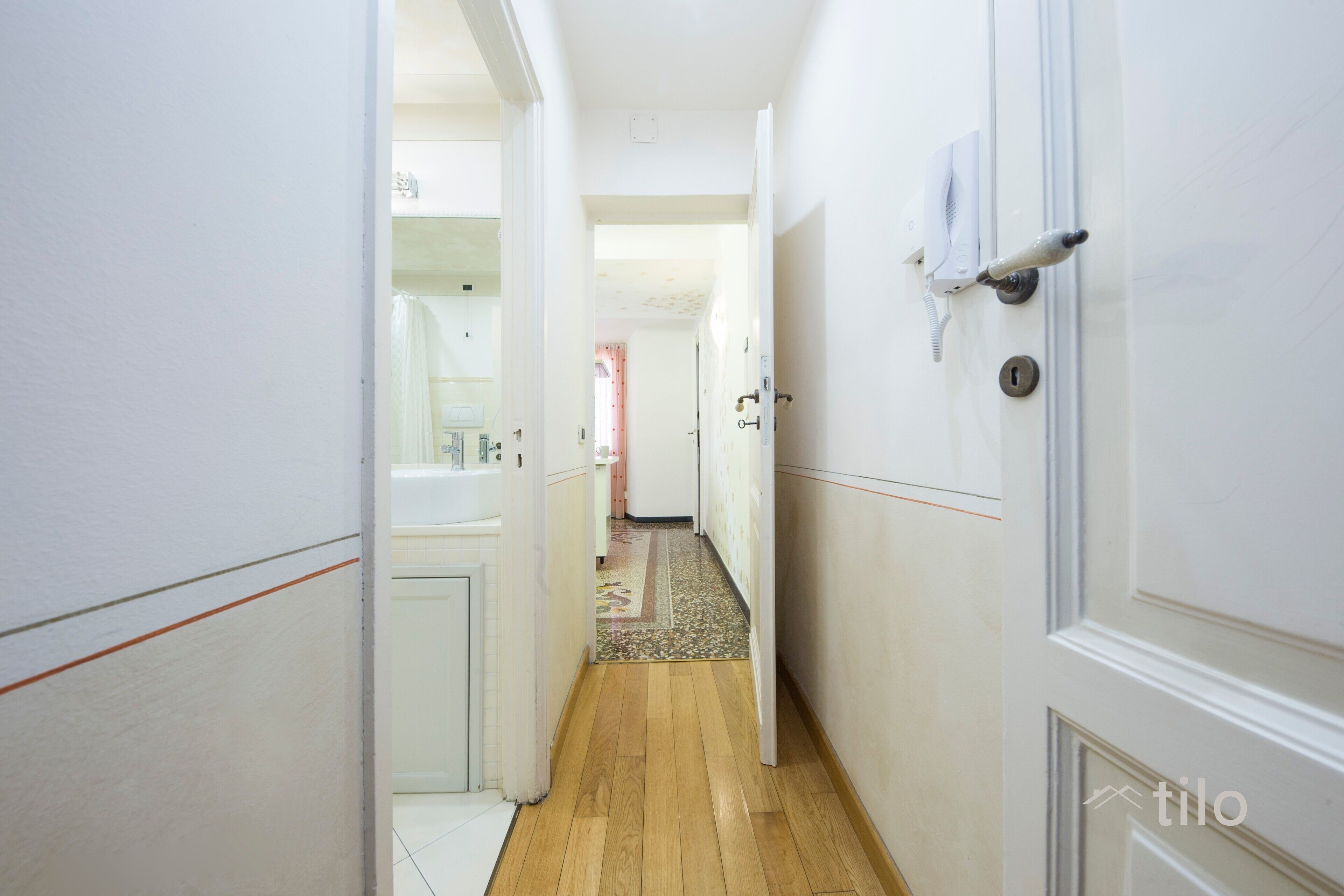 A narrow hallway with wooden flooring, white walls, and a door with a key lock.