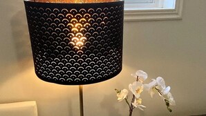 Eine Lampe mit schwarzem, perforiertem Schirm, ein kleiner Tisch mit einer Topforchidee und ein Fenster mit Blick auf ein Haus und Bäume.