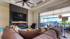 A modern living room with a large sofa, a flat-screen TV, and a view of a water body and outdoor seating area. - VRCC - Villa Taradise (Cape Coral)
