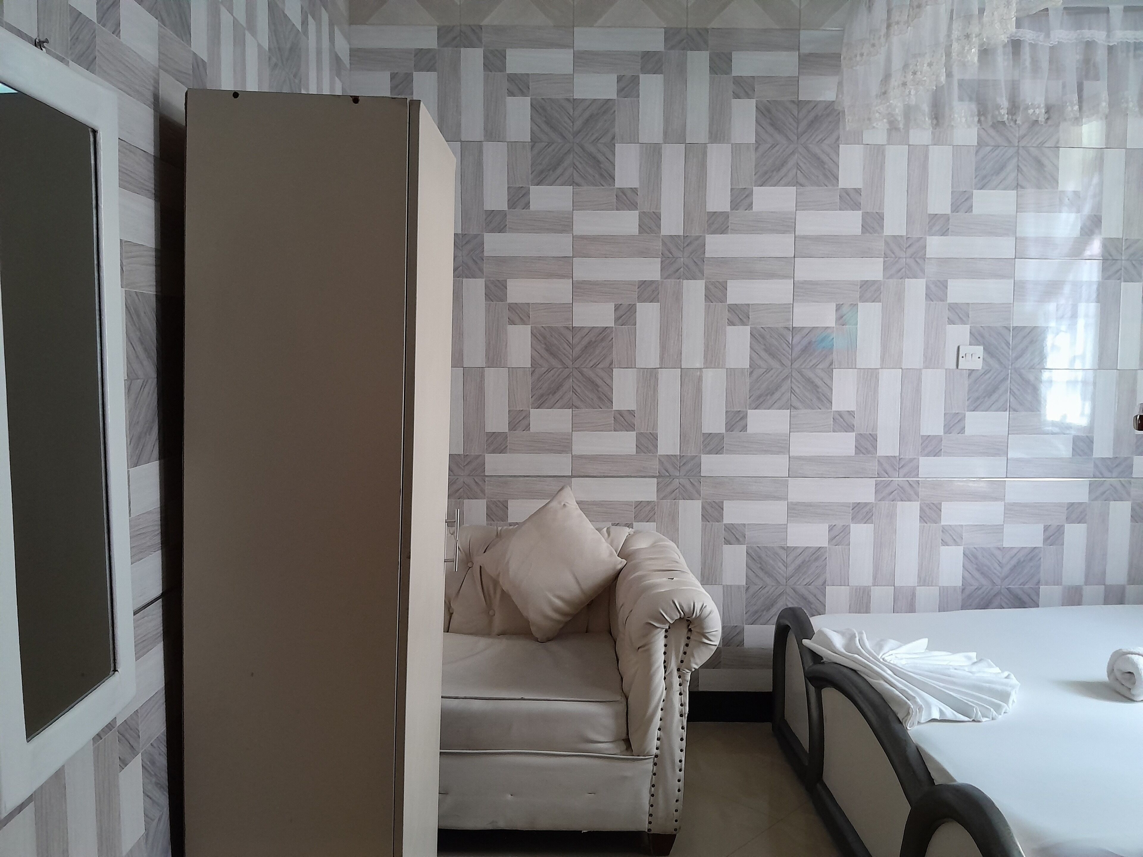 Quarto de hotel com parede de azulejos estampados, cama com colcha branca, uma cadeira e um guarda-roupa.