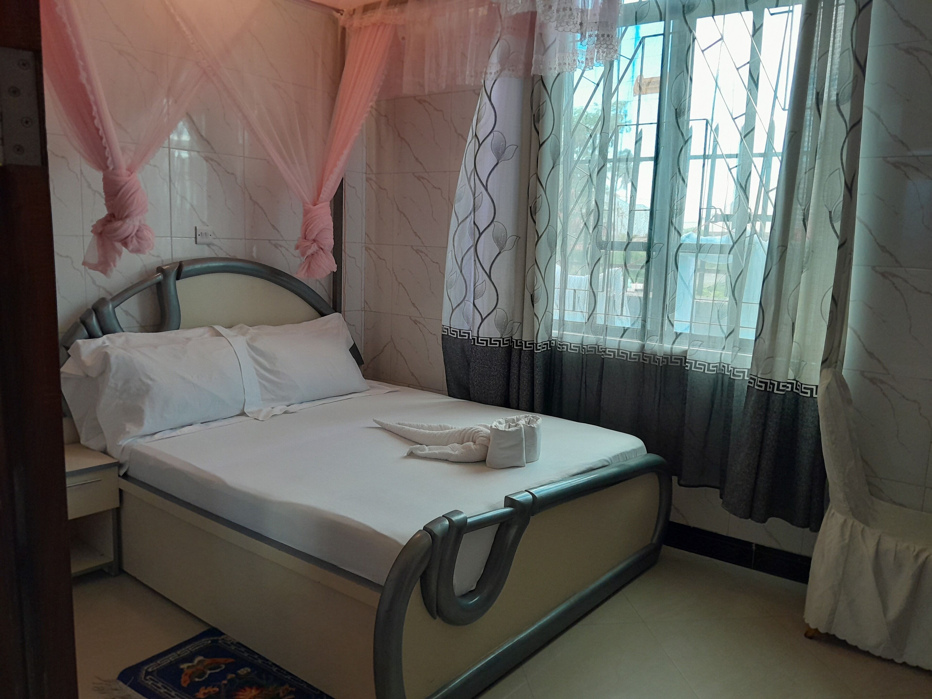 A neatly made bed with white linens and a single pillow, a bedside table with a lamp, and a window with patterned curtains.