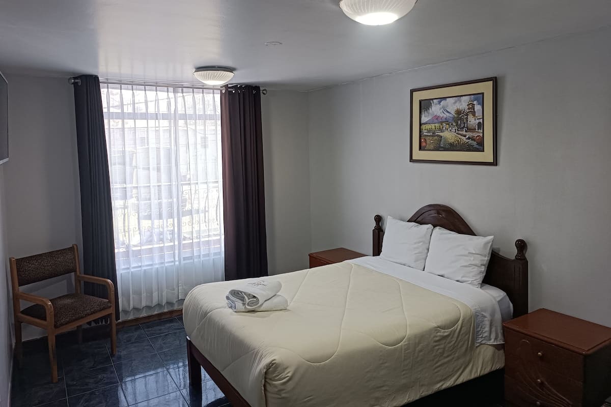 Ein Hotelzimmer mit Bett, Sessel, Nachttisch, einem Bild an der Wand und einem Fenster mit Vorhängen.