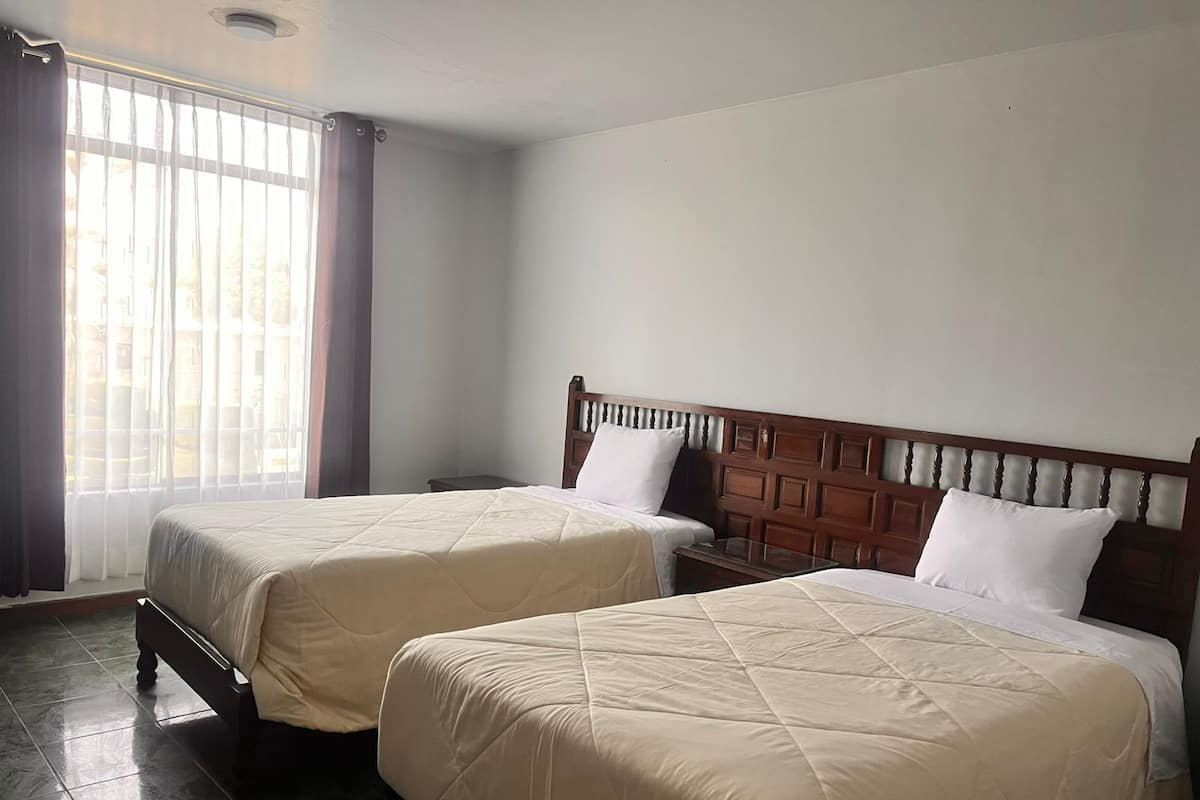 Ein Hotelzimmer mit zwei Betten, einem Holzkopfteil, einem Nachttisch und einem Fenster mit Vorhängen.