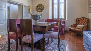 A dining room with a wooden table, chairs, and a decorative bowl. There is a window with lace curtains and a wall-mounted picture. - Orazio Liberty House,Lunigiana, Nature (Fivizzano)