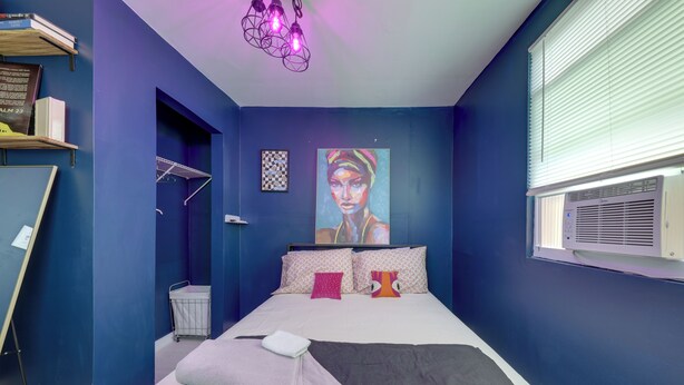 A bedroom with a bed, pillows, a painting, a window with blinds, and a wall-mounted air conditioner. - Pet-Friendly Fort Lauderdale Home: 8 Mi to Beach! (Fort Lauderdale)