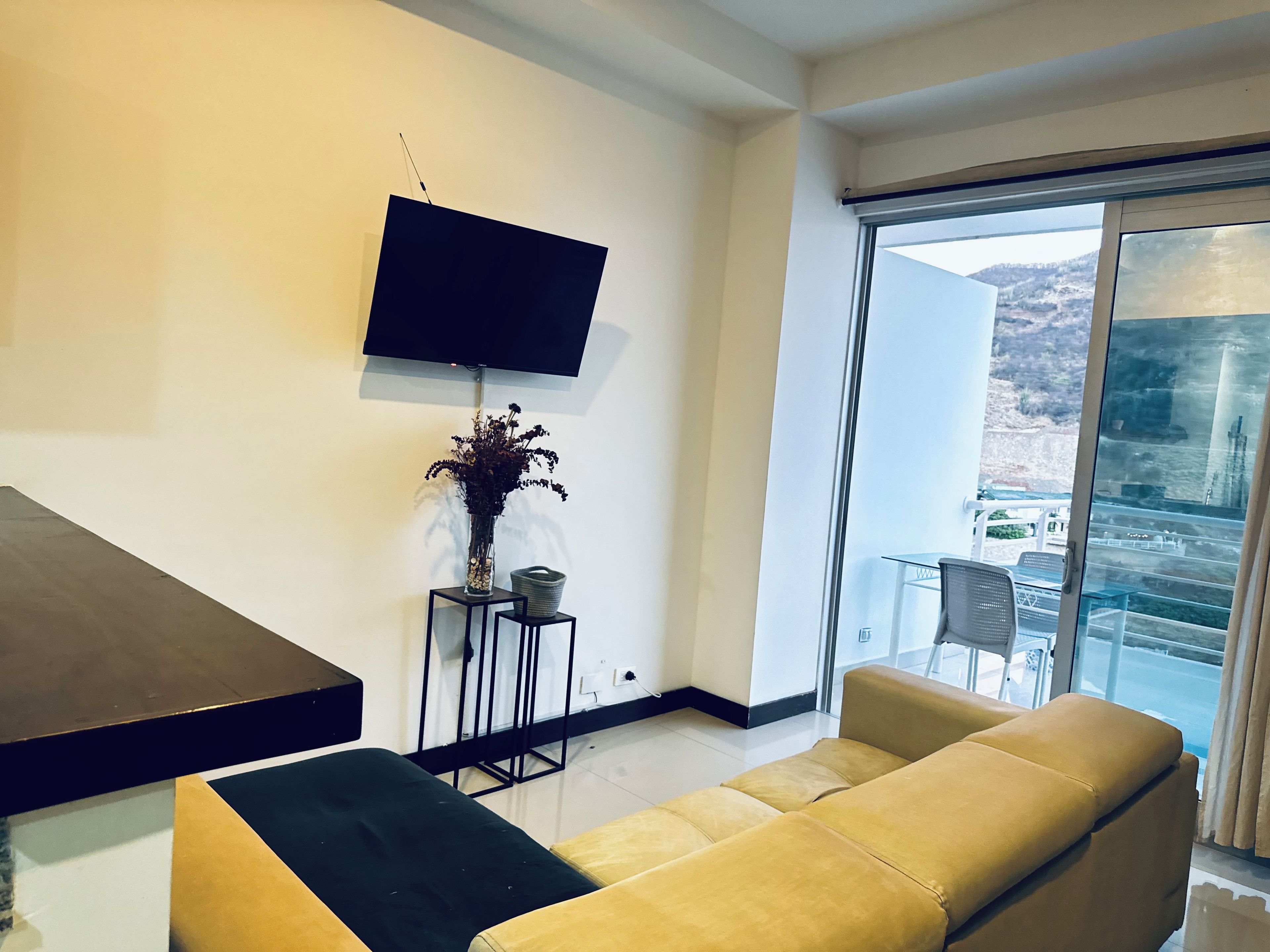A modern living room with a sofa, a flat-screen TV mounted on the wall, a small table with a vase, and a sliding glass door leading to a balcony.