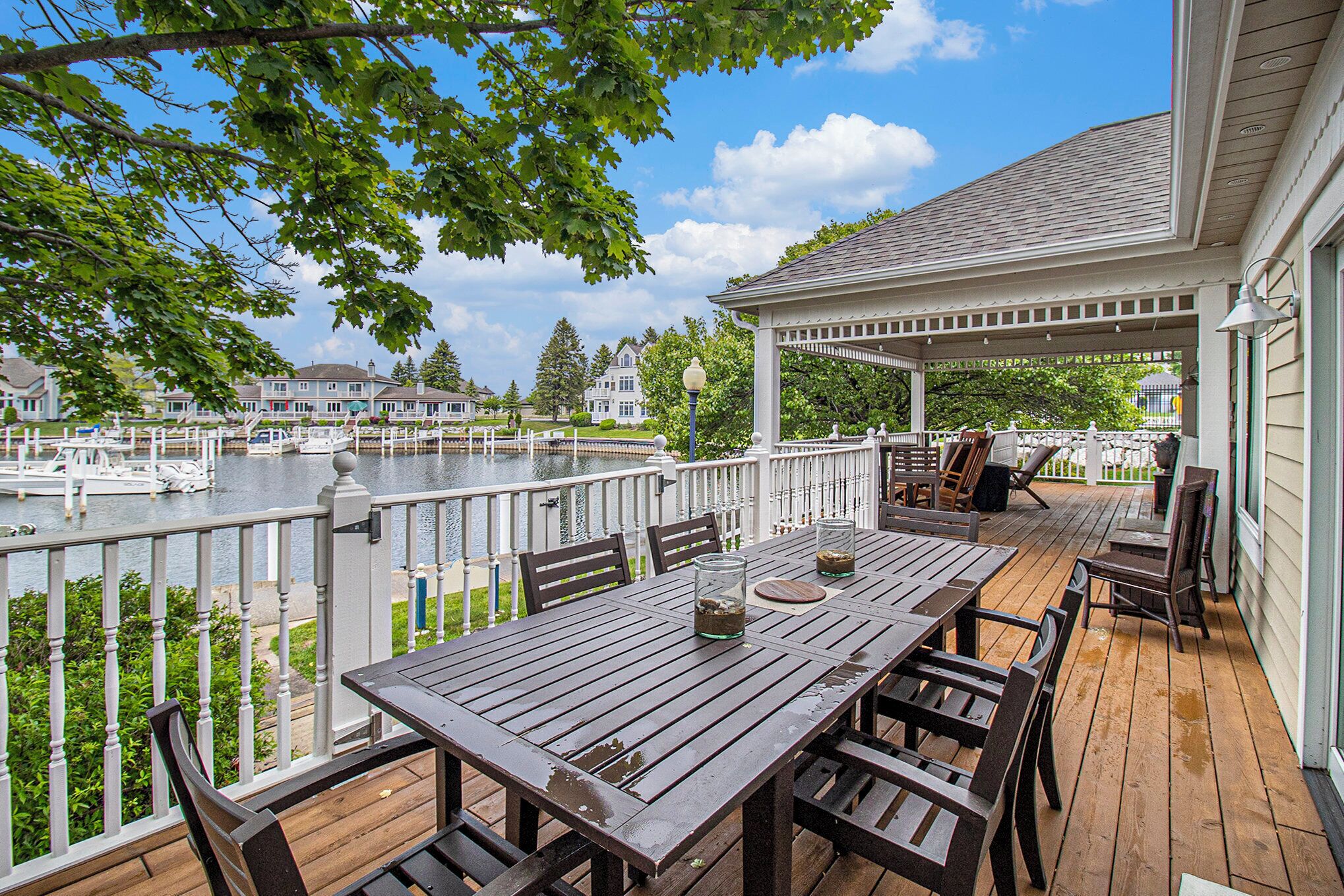 A wooden deck with a table and chairs, overlooking a body of water with boats and houses in the background.