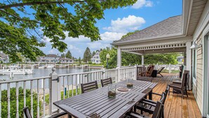 Eine Holzdielen-Terrasse mit Tisch und Stühlen, mit Blick auf ein Gewässer, im Hintergrund Schiffe und Häuser.