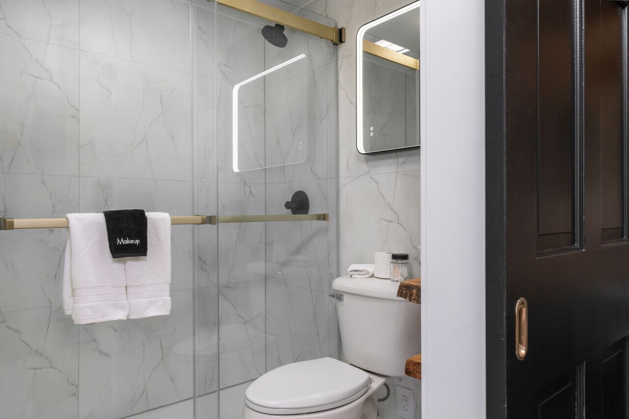 Een moderne badkamer met een marmeren wand, een wit toilet, een handdoekrek met een handdoek met de tekst 'Makeup', een spiegel en een douchehoek.