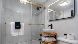Et moderne badeværelse med en brusekabine i glas, en hvid vask med en træhylde og et toilet med toiletpapir og et glas.