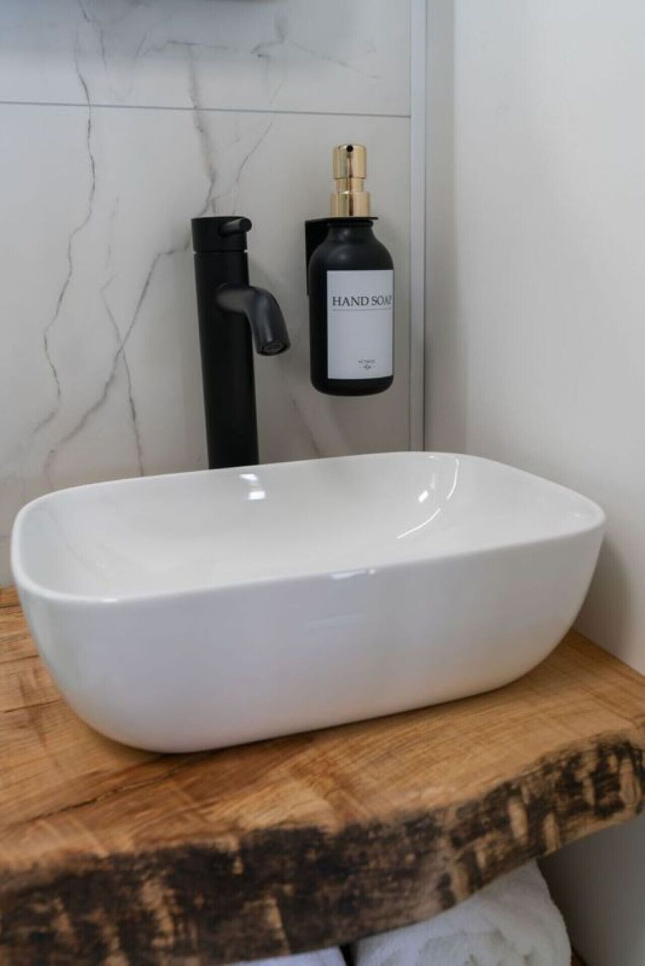 Un lavabo blanc, de forme arrondie, est posé sur un comptoir en bois, avec un robinet noir et un flacon de savon pour les mains à proximité.