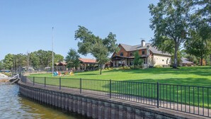 A riverside walkway with a stone bridge, a grassy lawn, and a large house with a red roof.