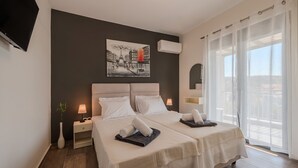 A hotel room with two beds, a painting on the wall, an air conditioning unit, a nightstand with a lamp, and a view of the outside through large windows. - 3A's Family luxury villa near Rethymno & all amenities (Rethymno Town)