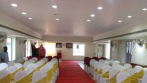 A conference room with rows of chairs covered in white and yellow sashes, a red carpet, and ceiling lights. - Sri sapthagiri lodge (Chirala)