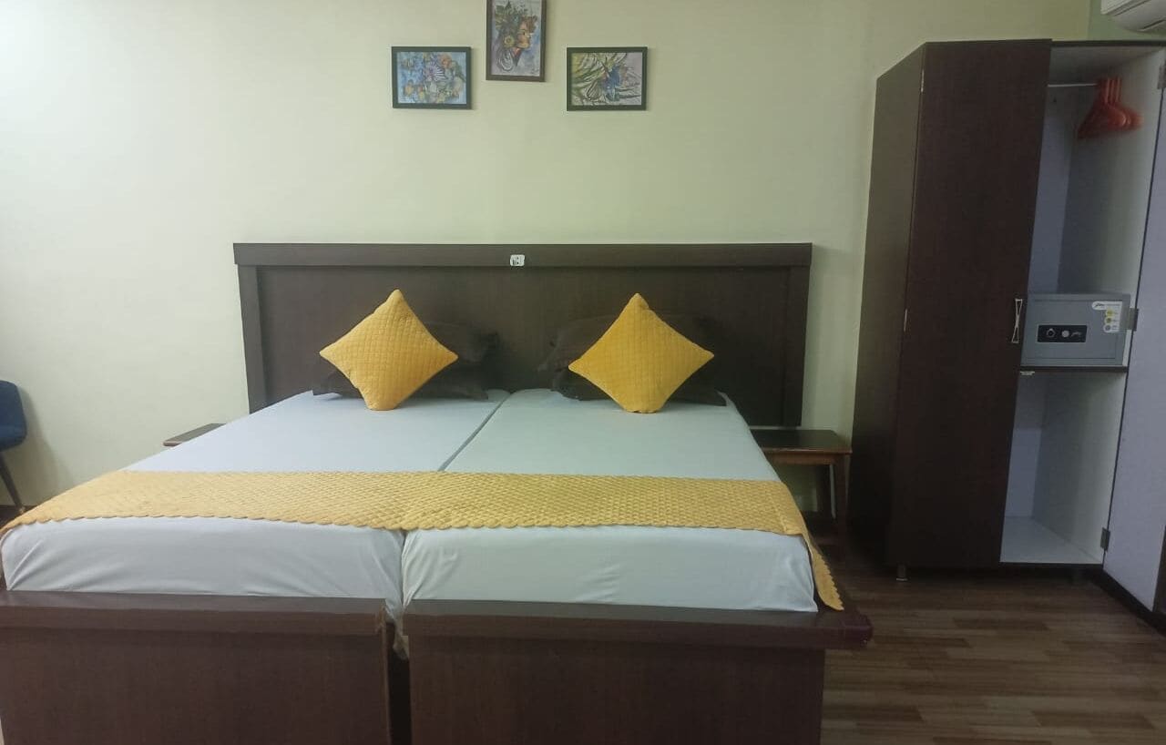 Uma cama de casal com cabeceira de madeira, duas almofadas, um cobertor amarelo e uma mesinha de cabeceira. Há três quadros na parede.