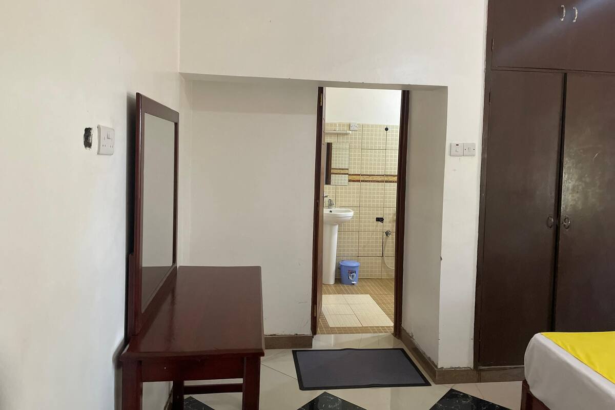Une pièce comprenant un lit, un bureau en bois, un miroir et une armoire. Une porte donne accès à une salle de bain équipée d’un lavabo et de toilettes.