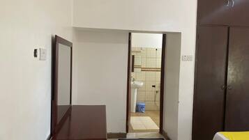 A room with a bed, a wooden desk, a mirror, and a wardrobe. There is a door leading to a bathroom with a sink and a toilet.