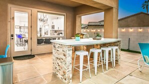 Un espace cuisine extérieur avec un comptoir-bar, un mur décoratif en pierre et une vue sur la maison en arrière-plan.