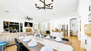 Un espace de repas moderne avec une longue table dressée pour six personnes, une cheminée et une cuisine en arrière-plan.