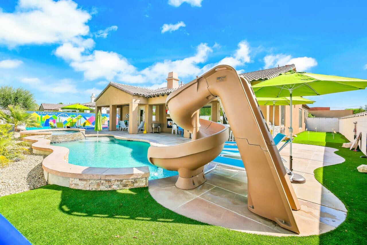 A swimming pool with a large slide, surrounded by a patio area with umbrellas and chairs.