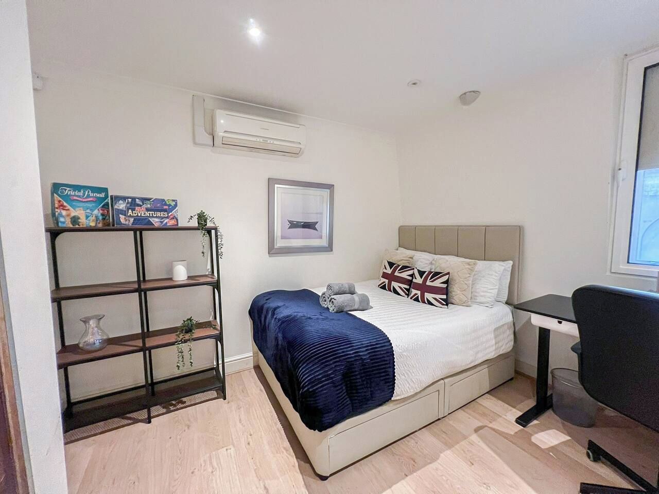 Un dormitorio con cama, escritorio, silla, estante con libros y un cuadro en la pared.