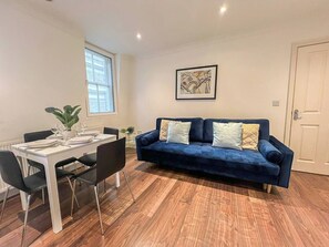 A modern living room with a dark blue sofa, a dining table set for four, and a large window providing natural light.