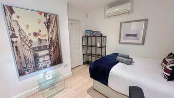 A modern bedroom with a bed, a glass table, a shelf with items, and a painting on the wall.