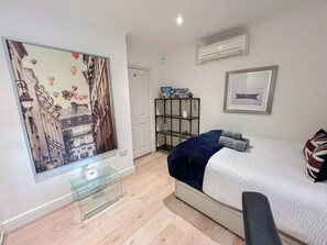 A modern bedroom with a bed, a glass table, a shelf with items, and a painting on the wall.