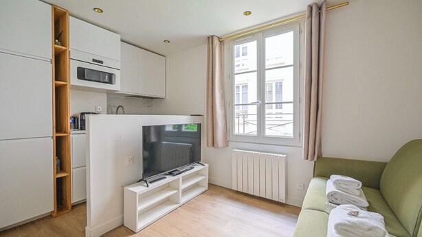 A compact living space with a kitchenette, a flat-screen TV, a sofa with towels, and a window with curtains.