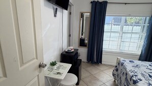 Una habitación pequeña y ordenada con una cama, un escritorio con una silla, un televisor montado en la pared y una ventana con cortinas.
