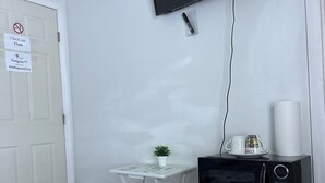 Una habitación pequeña con una puerta blanca, un taburete blanco, una mesita pequeña con una planta en maceta, un microondas negro y un televisor de pantalla plana montado en la pared.