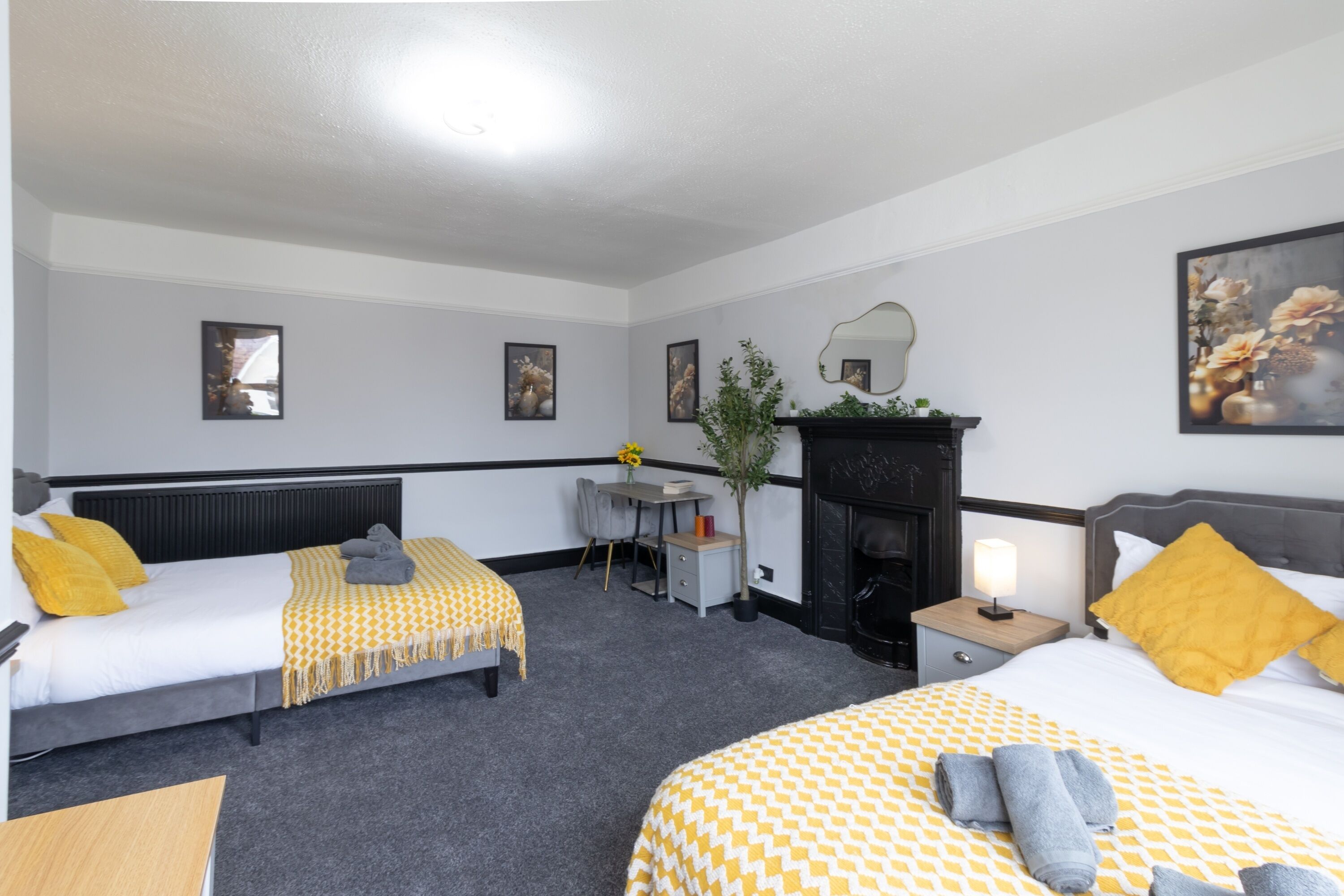 A hotel room with two beds, a desk, a chair, a fireplace, and framed artwork on the walls.