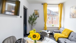 A modern living room with a dining area, featuring a sofa, a dining table set with plates, glasses, and a vase with sunflowers. - Modern 2-Bedroom Southsea Flat with Free Parking (Portsmouth)
