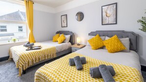 A hotel room with two beds, each with a patterned bedspread, pillows, and towels. There is a window with yellow curtains, a bedside table with a lamp, and wall art. - Modern 2-Bedroom Southsea Flat with Free Parking (Portsmouth)