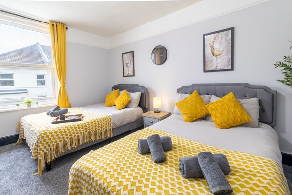 A hotel room with two beds, each with a patterned bedspread, pillows, and towels. There is a window with yellow curtains, a bedside table with a lamp, and wall art. - Modern 2-Bedroom Southsea Flat with Free Parking (Portsmouth)