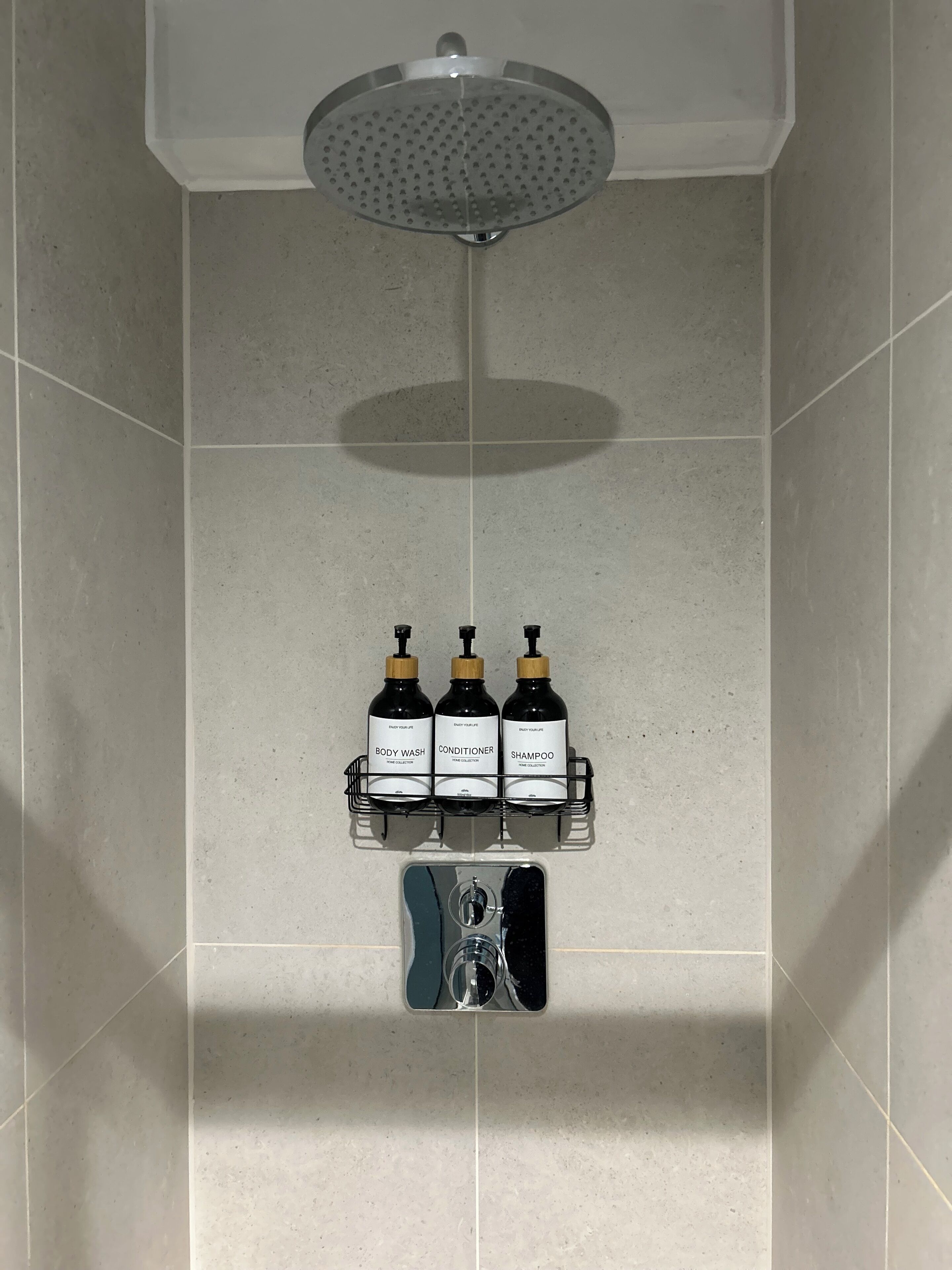 Zona de ducha con cabezal de ducha de lluvia, un estante con tres botellas que contienen gel de baño, acondicionador y champú, y un cabezal de ducha manual.
