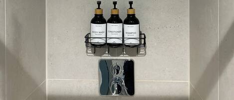 Zona de ducha con cabezal de ducha de lluvia, un estante con tres botellas que contienen gel de baño, acondicionador y champú, y un cabezal de ducha manual.