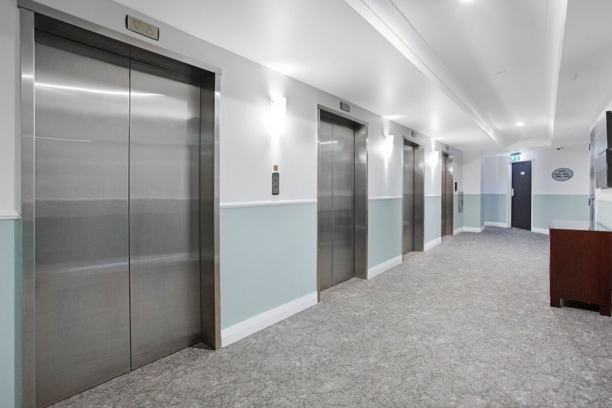 Corredor com quatro elevadores, piso coberto de carpete e um balcão de madeira à direita.