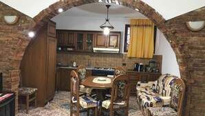 A kitchen with a brick archway, wooden cabinets, a dining table with chairs, and a sofa. - Through Mangalem (Berat)