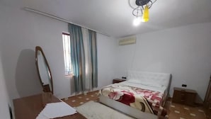 A bedroom with a bed, a wooden dresser, a mirror, a window with curtains, and a ceiling light. - Through Mangalem (Berat)