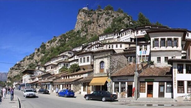 A street view of a town with buildings, a mosque, and a mountain in the background. - Through Mangalem (Berat)