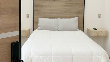 Una cama bien hecha con cabecera de madera, una mesita de noche con control remoto y un interruptor de luz en la pared.