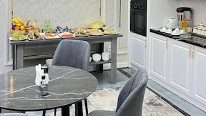 A modern kitchen with white cabinetry, a dining table with a marble top, and a breakfast buffet with various foods. - Annur Boutique Hotel (Samarkand)