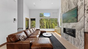 A modern living room with a leather sectional sofa, a round coffee table, a fireplace, and a large window offering a forest view. - Luxury Townhome, Steps From Ski Lifts w/ Private Hot Tub & Mountain Views! (Keystone)