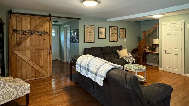 A living room with a dark-colored sofa, a patterned bench, a small round table, and a wooden door with a 'Welcome to Our Home' sign. - Northern Maine Getaway-Direct Trail Access (Fort Fairfield)