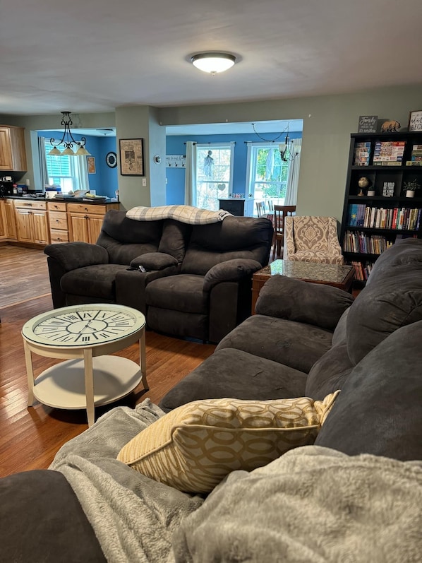 A living room with a dark brown sofa, a round table with a clock, and a bookshelf. - Northern Maine Getaway-Direct Trail Access (Fort Fairfield)