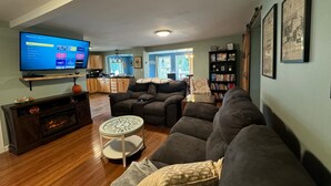 A living room with a flat-screen TV, a fireplace, a sofa, a coffee table, and a bookshelf. - Northern Maine Getaway-Direct Trail Access (Fort Fairfield)