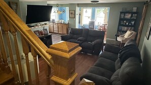 A living room with a wooden staircase, a flat-screen TV, a sofa, and a bookshelf. - Northern Maine Getaway-Direct Trail Access (Fort Fairfield)